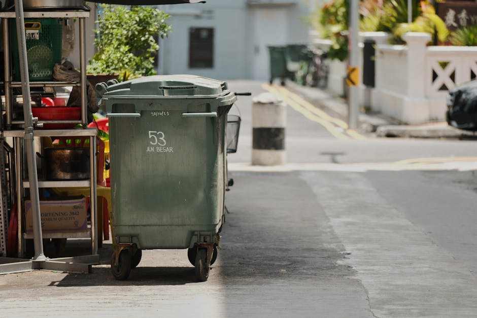 A green wheeled rubbish bin with a flat lid, positioned on a sidewalk near a storage rack containing various household items such as plastic containers, and located in an urban residential area. The b